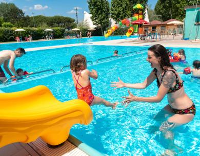 Bambini giocano in piscina con scivolo giallo e adulti.