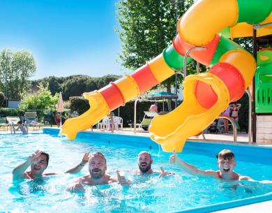 Four men smiling in a pool with a colorful slide.