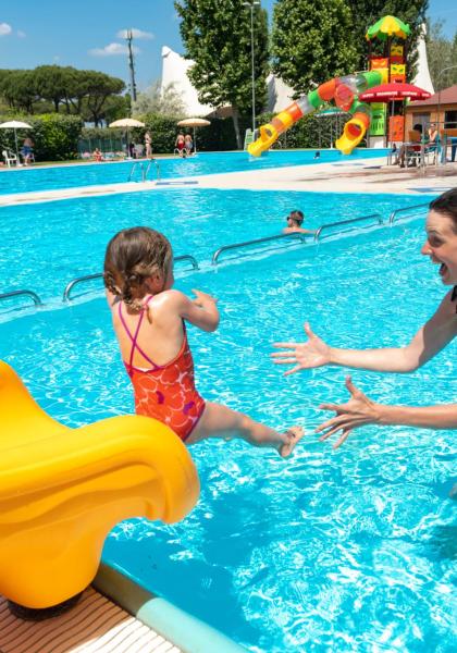 Bambini giocano in piscina con scivolo giallo e adulti.