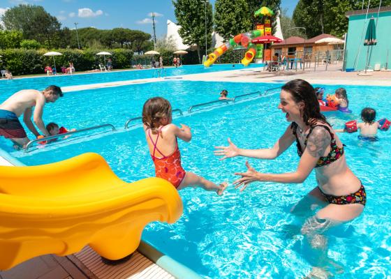 Bambini giocano in piscina con scivolo giallo e adulti.