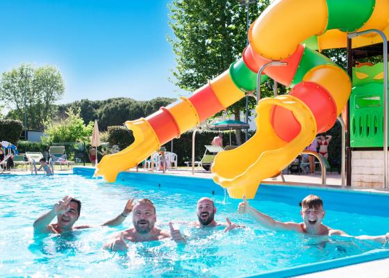 Quattro uomini sorridenti in piscina con scivolo colorato.